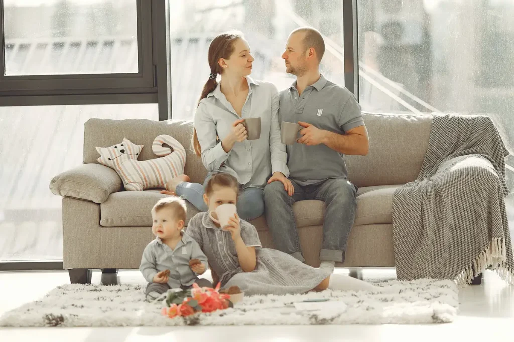 Parents sitting on a couch drinking coffee while kids play on the living room rug